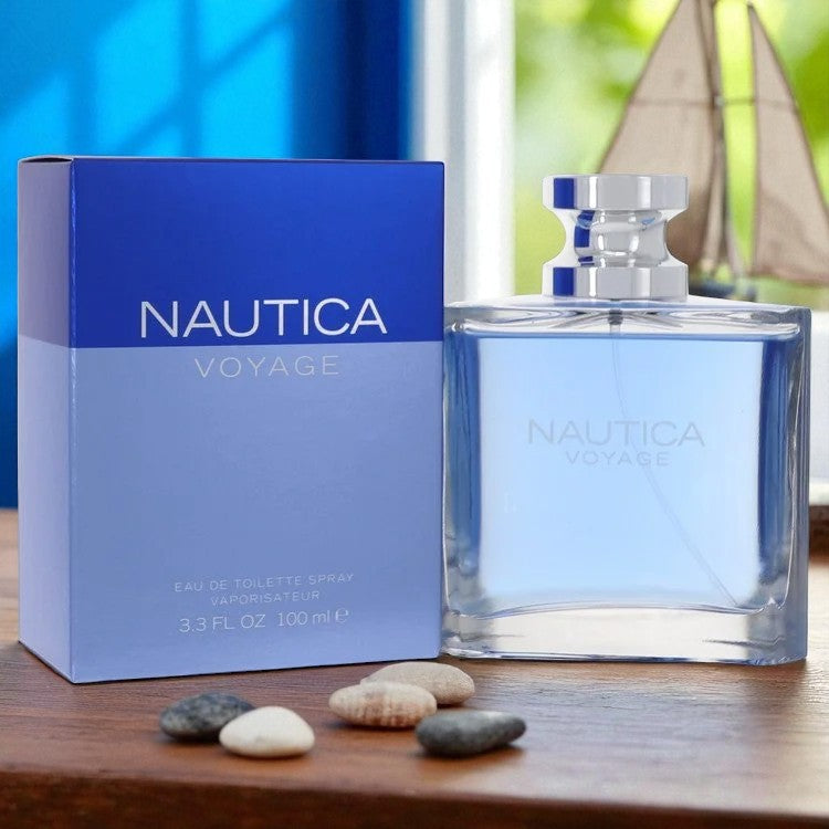 Nautica Voyage Cologne for Men in a sleek blue 3.3 oz bottle, placed next to its matching box on a wooden surface with smooth stones and a sailboat in the background, evoking a fresh, aquatic vibe inspired by the sea.