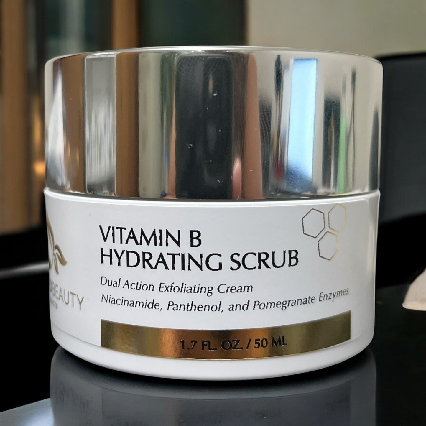 High-resolution close-up of Reiland Vitamin B Hydrating Scrub jar placed on a glossy surface with ambient light reflecting from the silver lid.