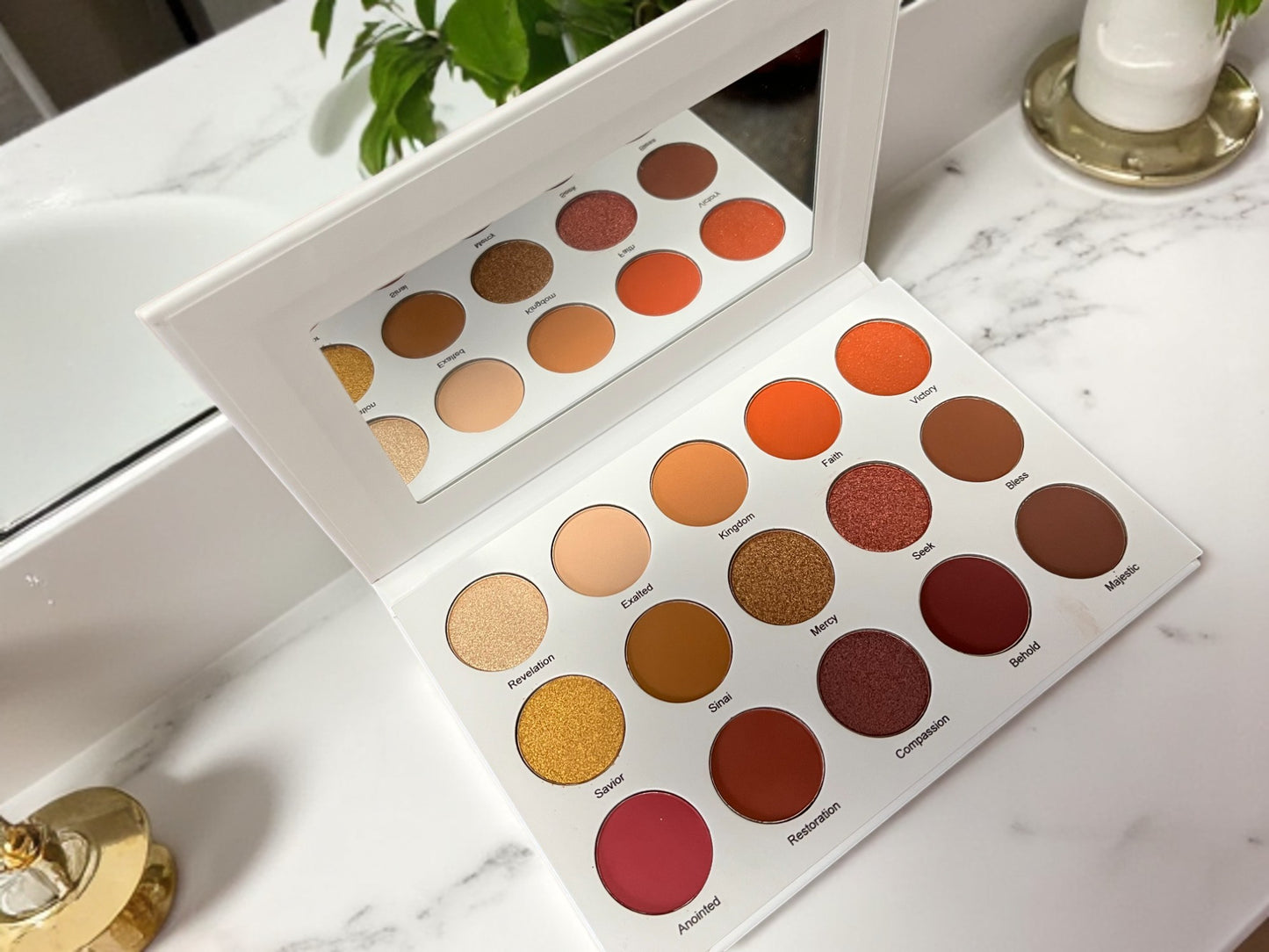 Open Velvet Bloom 15-color eyeshadow palette by Reiland Beauty Products with built-in mirror, showcasing matte and shimmer shades on a white marble vanity with gold and green plant accents.