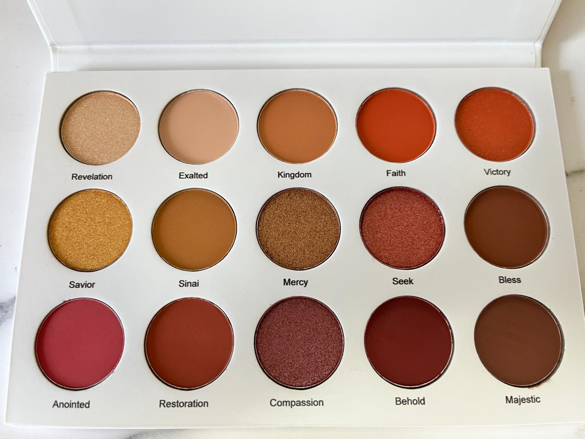 Flat lay of Reiland Beauty Products' Velvet Bloom 15-color eyeshadow palette showing matte and shimmer shades with inspirational names like Revelation, Faith, Mercy, and Anointed on a white vanity background.