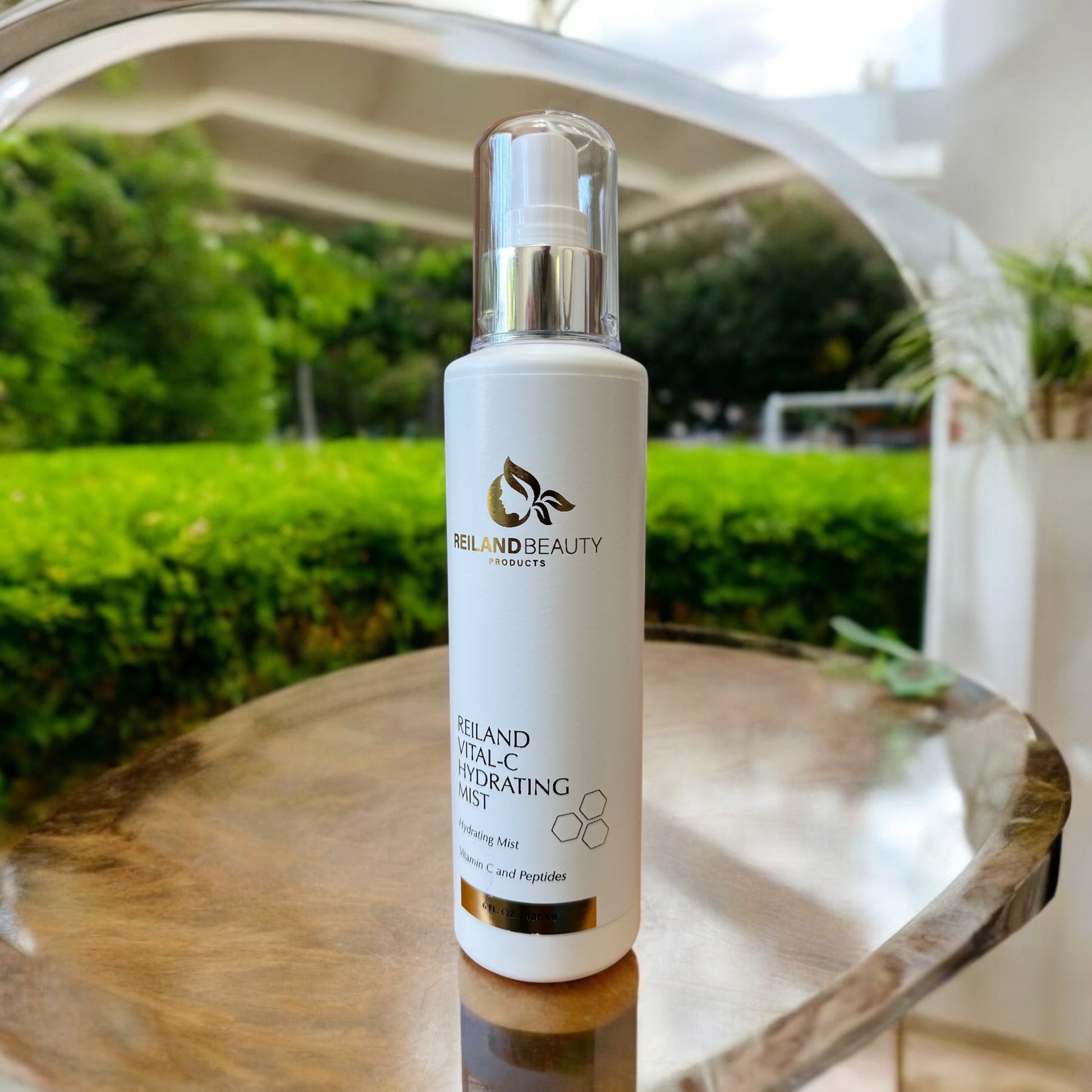 Reiland Vital-C Hydrating Mist – Vitamin C face spray for hydration, collagen boost, and skin refreshment. Luxury skincare mist on a reflective outdoor table with greenery in the background.